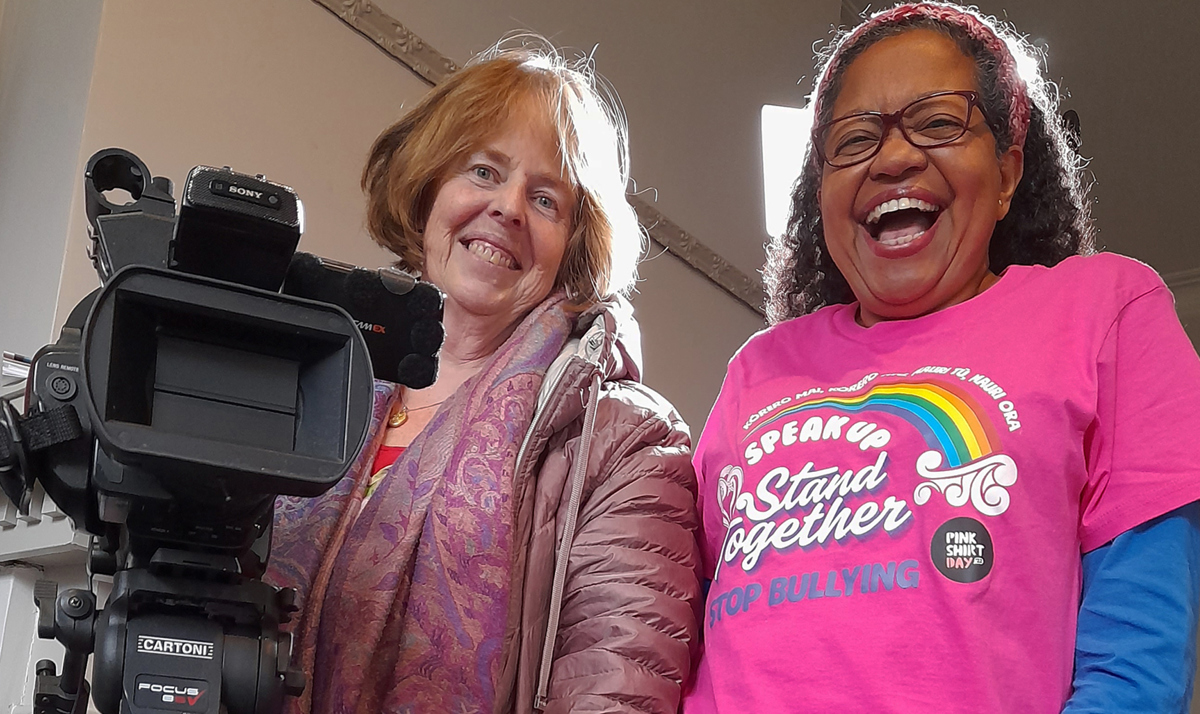 Two women involved in a documentary stand smiling beside a film camera